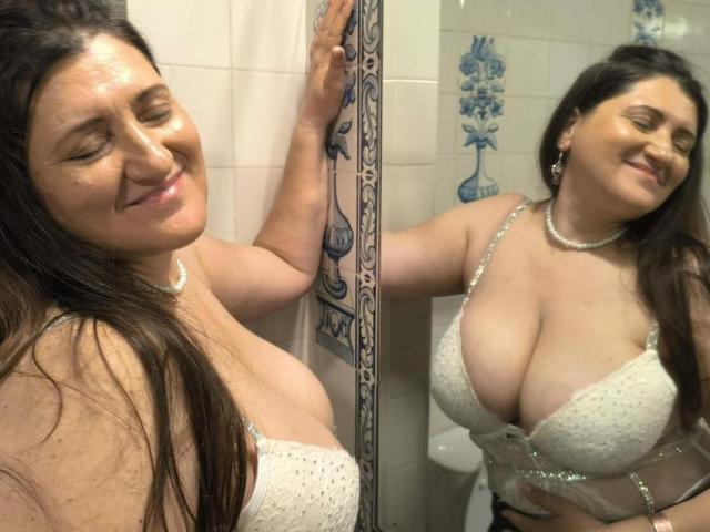 BlissSophia, a curvy woman with large tits, smiling in a bathroom, wearing a white, lace, sheer bra, reflected in a mirror, brown hair, pearl necklace, eyes closed.