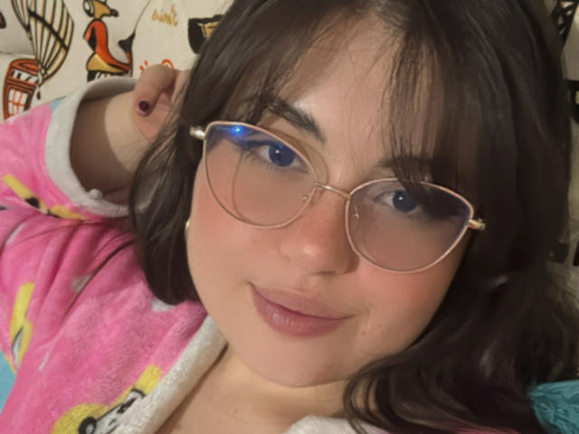 VicluX with glasses, pink pajamas, and black hair, looking at camera with a slight smile. She's lying down with her arm raised, showing a pink nail.