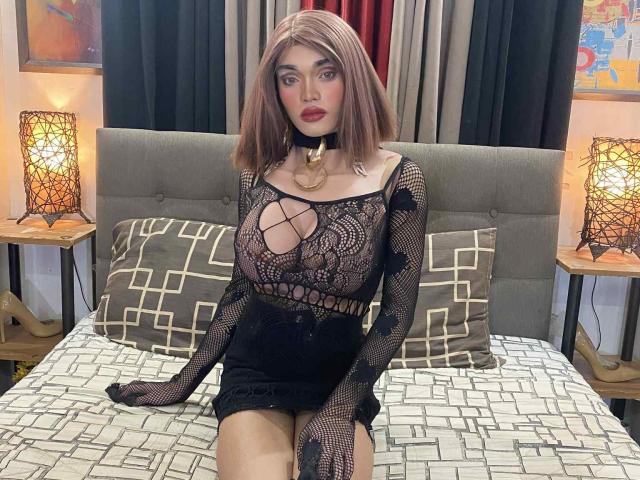 HorseCockKharlaTS with brown bob, red lipstick, black lace dress, big tits, choker, black skirt, sitting on bed with geometric pattern, lamps, and curtains in the background.