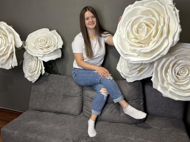 MavkaCute, avec des seins, assise sur un canapé gris, portant un jean déchiré, des chaussettes blanches et un t-shirt blanc, touchant une grande rose blanche, souriante, cheveux bruns, peau claire, sol en bois..