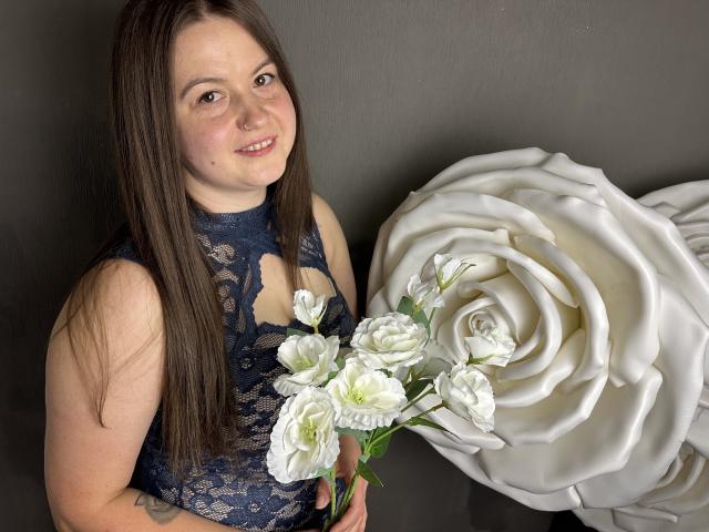 Mavka: Een meisje met bruin haar, gekleed in een kantjurk, die witte bloemen vasthoudt, naast een gigantische witte roos, en lacht naar de camera. Haar borsten zijn klein en haar achterkant is niet zichtbaar..