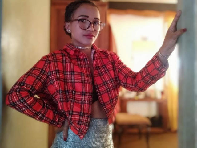 Tyahlive with glasses, red plaid shirt, small tits, choker, and grey high-waisted pants, standing against wall, hand on wall, smiling, brown hair in buns.
