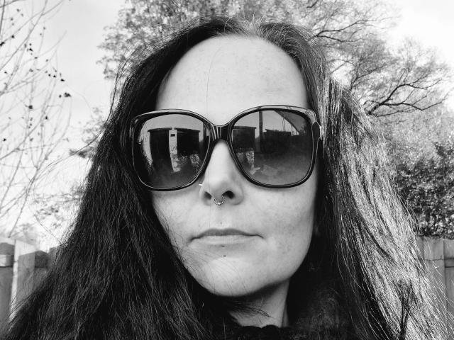 NoireEtoile-hot has long dark hair, a nose ring, and is wearing large black sunglasses, with a subtle smirk, in a black-and-white selfie. Her reflection is in the sunglasses.
