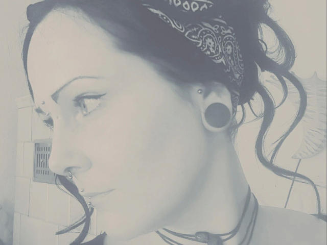 NoirEtoile-hot's face in a black-and-white photo, showing a nose ring, bandana, large ear gauges, and a septum piercing, with wavy hair and a neutral expression.