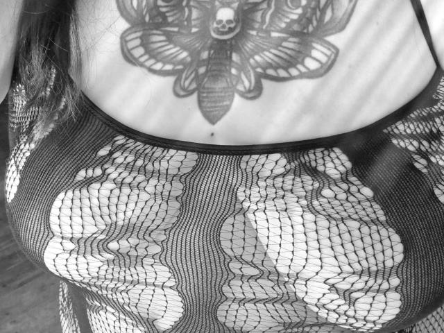 Noiretoile-hot's tits in a black mesh top, revealing wavy patterns. Upper chest tattoo of a detailed moth with a skull in the center. Black and white photo.