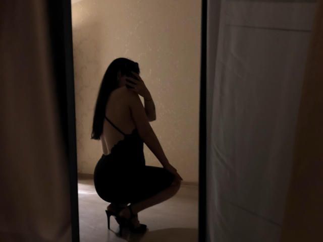 PannochkaX está en cuclillas, vestida con lencería negra y tacones altos, sosteniendo un teléfono frente a su cara, y su reflejo se ve en un espejo. Su trasero es prominente y tiene el pelo largo y negro..