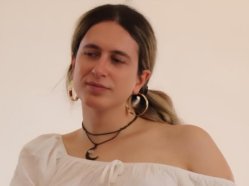 NonbinaryBerry ha la pelle chiara, gli occhi marroni e un leggero sorriso, indossa una maglietta bianca con una spalla scoperta, una collana nera con mezzelune e grandi orecchini a cerchio dorati..