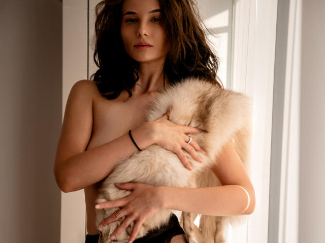 AmiraBlush with wavy brown hair, topless, holding a fluffy white fur, covering her tits with her hands, wearing a black bracelet, ring on her right hand, sunlight.