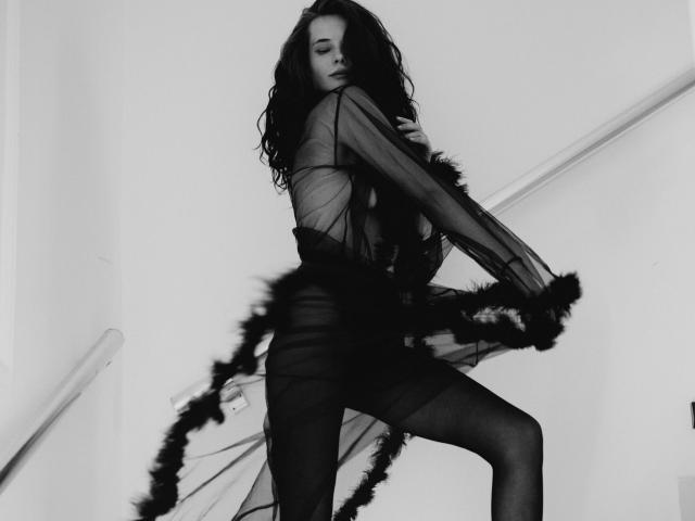 AmiraBlush with dark wavy hair, wearing sheer black dress with feathers, standing on stairs, black stockings, holding dress, looking seductively over shoulder, black-and-white photo.