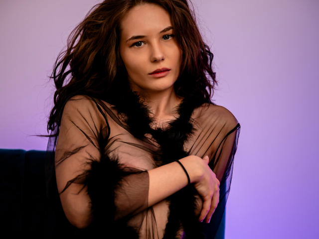 AmiraBlush with wavy brown hair, wearing a sheer black dress with black feather accents, arms crossed, looking at camera with a slight smile, soft purple lighting.