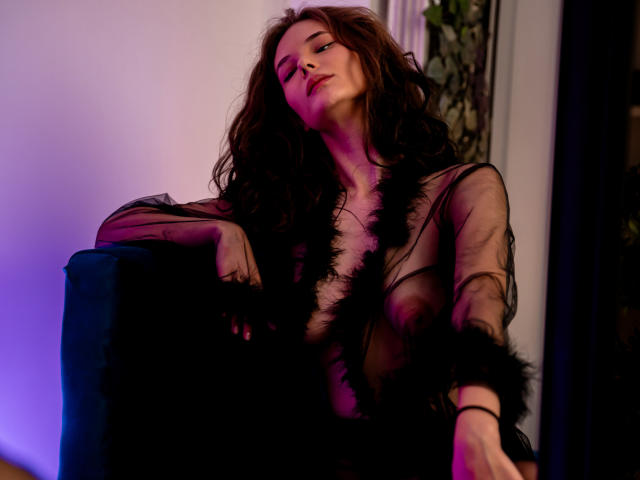 AmiraBlush with wavy brown hair, wearing a sheer black robe, exposing her tits, and black feather accents, leaning back in a blue chair, eyes closed, under purple and blue light.