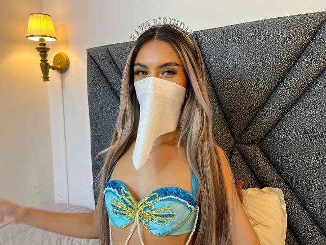 DalilaAliX with long blonde hair, blue lace-up bra, yellow embroidery, pearl chains, white mask, wearing a "bride" headpiece, standing in front of a gray, textured headboard.
