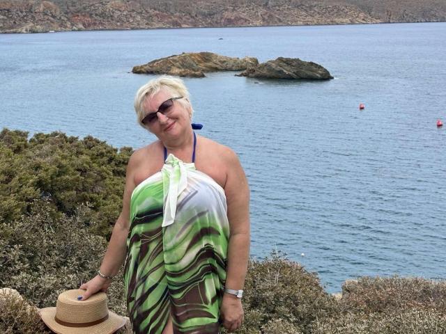 MadyAllav with short blonde hair, wearing sunglasses, a green and white striped halter dress, holding a straw hat, standing by a rocky shoreline with a blue sea.