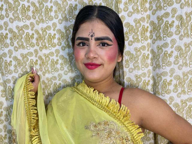 NayaYalitza with dark hair, red lipstick, and a yellow saree with gold embroidery, holding the edge. She's got a bindi on her forehead, smiling with pink blush.