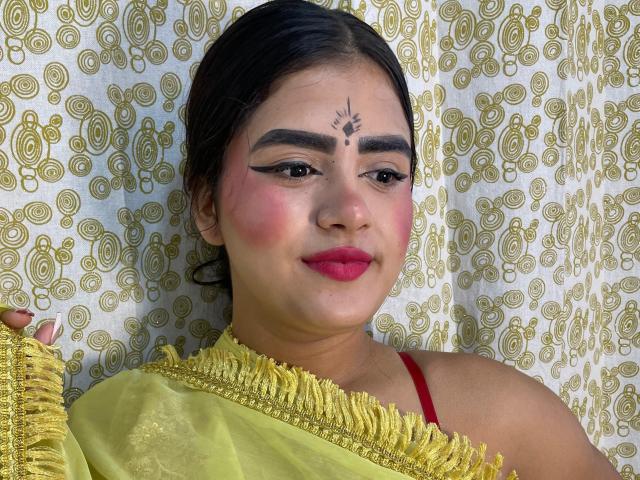 NayaYalitza with dark hair, brown eyes, and pink cheeks, wearing a yellow cloth, red strap, and a bindi, smiling with pink lipstick, holding yellow fabric.