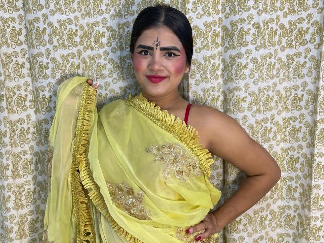 NayaYalitza with medium brown skin, black hair, red lipstick, wearing a yellow saree with gold embroidery, holding the saree's edge, smiling, red bra visible, decorated forehead dot.