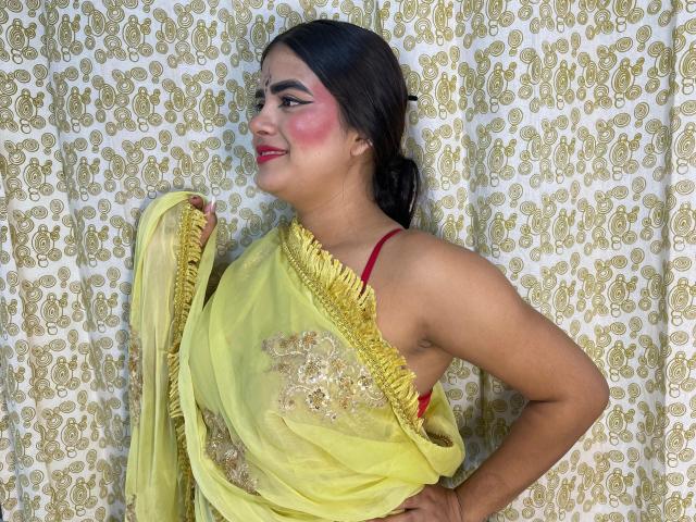 NayaYalitza with yellow saree, red bra visible, dark hair in ponytail, pink blush, smiling, holding saree, gold patterned background, her tits slightly showing through sheer fabric.
