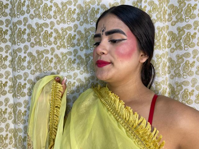 NayaYalitza with yellow saree, holding it up, showing red strap, pink cheeks, black eyebrows, red lipstick, and a bindi on her forehead. She has dark hair and a slight smile.
