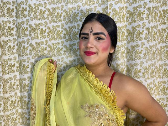 NayaYalitza, dark-haired woman, wearing yellow saree with gold embroidery, red bra visible, makeup with pink blush, forehead bindi, holding saree, smiling, medium skin, black eyebrows.