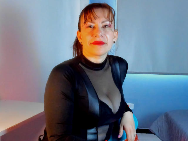 AmaraMoon with medium tits, wearing a sheer black top, seated, looking at camera, red lipstick, brown hair, slight smile, orange-blue light, right hand on chair.