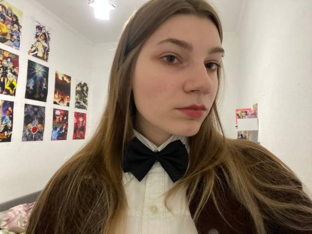 PureRadiance has long brown hair, a black bowtie, and a brown cardigan over a white shirt. Her face shows a slight pout, and she's looking into the camera.