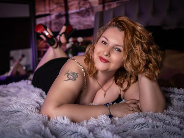 EmmaaPark with wavy red hair, black dress, and gold necklace, laying on a fluffy gray bed, showing a tattoo on her right arm, smiling at the camera.