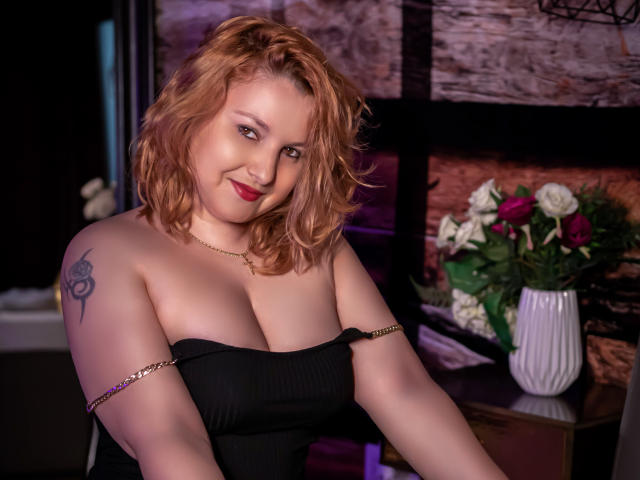 EmmaaPark with curly red hair, black strapless top, gold chain, tattoo on left arm, red lipstick, smiling, large tits, necklace, bouquet of roses in background, dim lighting.