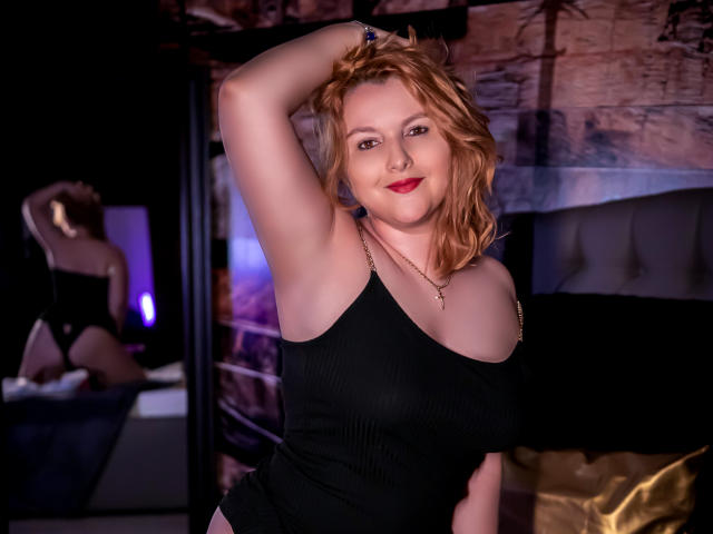 EmmaaPark with red hair, black ribbed tank top, raising arm, showing tits, smiling, mirror reflecting her ass, purple and blue lights, black and gold bed, necklace.