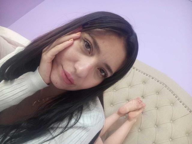 CattsyCathy with long black hair, light skin, and brown eyes, smiling softly, resting her hand on her cheek, wearing a white ribbed top, feet up, showing a cute pose.
