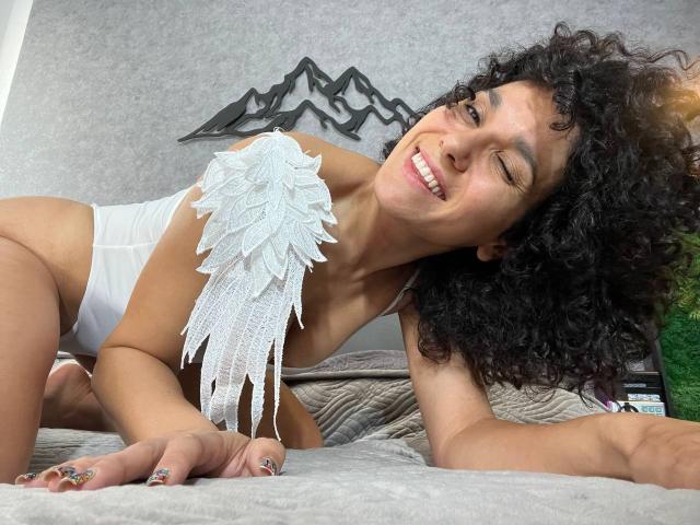 CurliestSue, with curly black hair and tan skin, winks, lying on a bed in white underwear, featuring a white lace wing on her right shoulder.