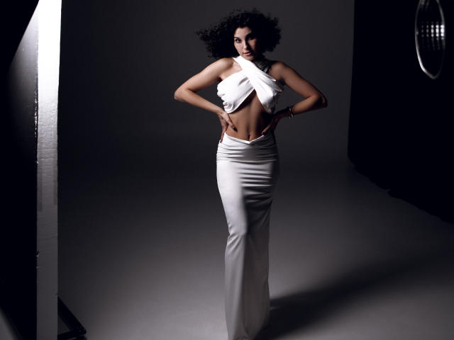 CurliestSue with curly black hair, wearing a white, halter-style, high-cut dress, hands on hips, exposing her toned abs and small, perky tits. She's standing in a dark studio.