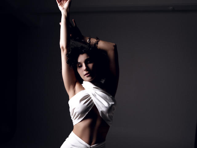 CurliestSue with dark curly hair, arms raised, wearing a white wrap-around dress, exposing her toned midriff and small, perky tits, standing in dim light.