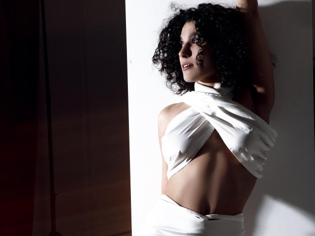 CurliestSue with curly black hair, wearing a white halter dress, exposing her back and small tits, stands in bright sunlight, arm raised, showing a sexy, confident smile.