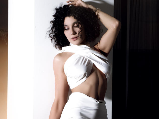 CurliestSue with curly black hair, wearing a white, criss-cross, halter-top dress, exposing her tits and abs, standing with one arm above her head, looking down.