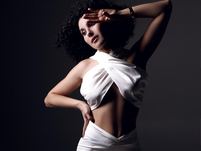 CurliestSue with curly black hair, wearing a white halter dress that exposes her tits and a sliver of her ass, posing with one arm above her head.