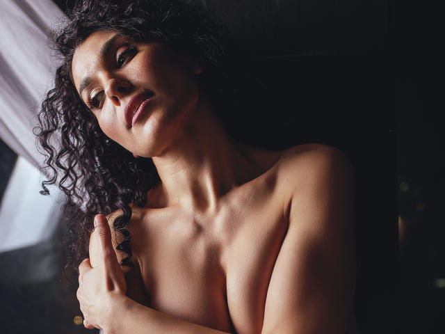 CurliestSue with curly black hair, medium brown skin, and small, perky tits, crosses arms, smiling softly, standing topless in dim light, showing a sexy, confident, and relaxed vibe.
