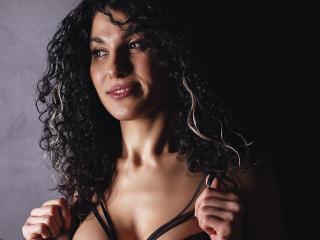 CurliestSue has curly black hair with white highlights, holding black straps, smiling with pink lips, brown eyes, and a light brown skin tone, showing small tits, in a dark setting.