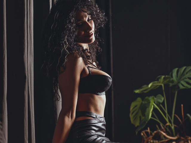 CurliestSue with curly black hair, wearing a black leather bra and matching skirt, standing in dim light, showing her toned abs and small, firm tits. Green plant on right.