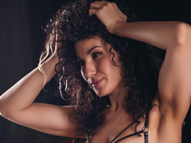 CurliestSue with curly black hair, smiling, arms raised, showing armpits, wearing a black strap top, dark background, soft light, confident, attractive, brown skin, slight smile.
