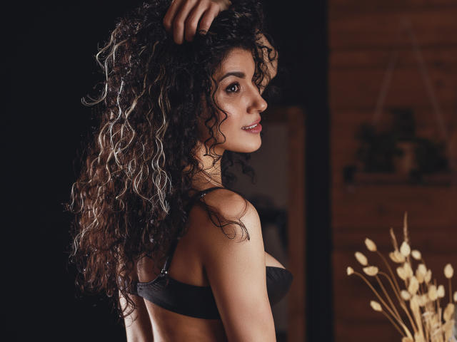 CurliestSue with curly black hair, smiling, wearing black bra, side profile, hand in hair, medium tits, brown eyes, soft lighting, wooden wall, dried plant in background.