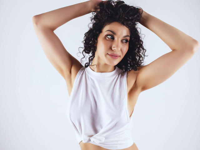 CurliestSue with curly black hair, arms raised, wearing a white sleeveless top tied at the front, exposing her tits and armpits, smiling slightly, against a plain white background.