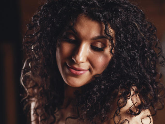 CurliestSue has dark curly hair, closed eyes, and a soft smile, with subtle makeup and warm skin. She's looking down, showing her pretty nose and full lips.