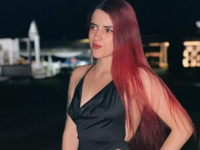 LaiaJonnes with long red hair, wearing a black dress, showing her tits in the photo, standing at night with a slight smile. Her ass is not visible.