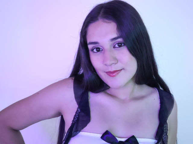 NinaClare with long black hair, wearing a strapless white top with black ribbons, smiling with pink lipstick, blue-tinted skin, and dark eyeliner. Her tits are small and cute.
