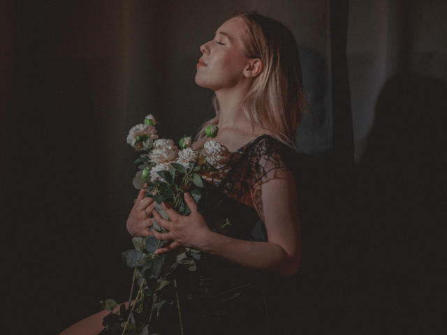 LanaRoselle, met blond haar, met gesloten ogen, die witte en groene bloemen vasthoudt, gekleed in een zwarte kanten jurk, zittend in een donkere, schaduwrijke kamer, met een vredige en ontspannen uitstraling..