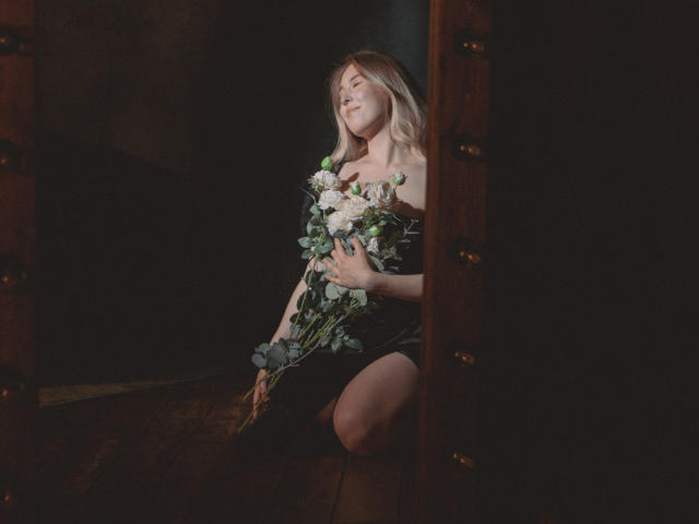 LanaRoselle è inginocchiata in una stanza buia, tiene in mano un bouquet di fiori bianchi e verdi, con gli occhi chiusi e un sorriso dolce, indossando un abito nero..