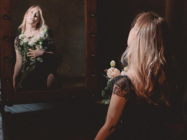 LanaRoselle mit blonden Haaren, die in einen Spiegel schaut, Blumen hält, ein schwarzes Spitzenkleid trägt, spiegelt sich in einem hölzernen Spiegel wider, dunkler Raum, weiches Licht, dezentes Tattoo am rechten Arm..