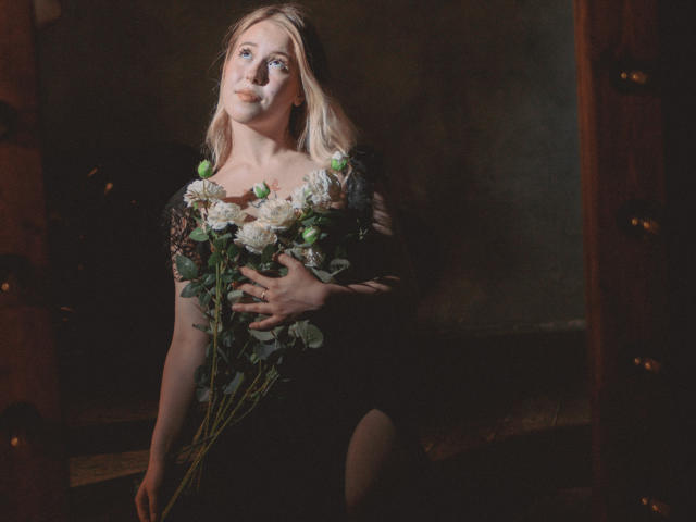 LanaRoselle mit platinblondem Haar, die weiße Rosen und grüne Knospen hält, schwarze Dessous trägt, nach oben schaut und ein sanftes Lächeln hat, in gedämpftem, warmem Licht, wobei ihre Brüste und ihr Hintern zu sehen sind..