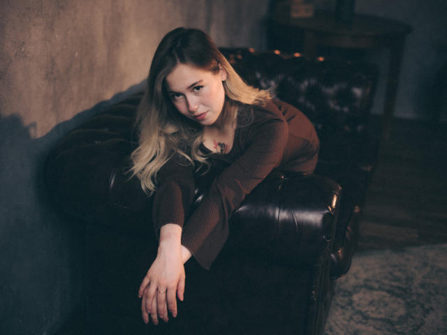 LanaRoselle leunt tegen een donkerbruine leren bank, met haar armen gekruist, en draagt een bruine ribgebreide top. Ze heeft blond haar en een subtiele glimlach, en kijkt op met een verleidelijke blik..