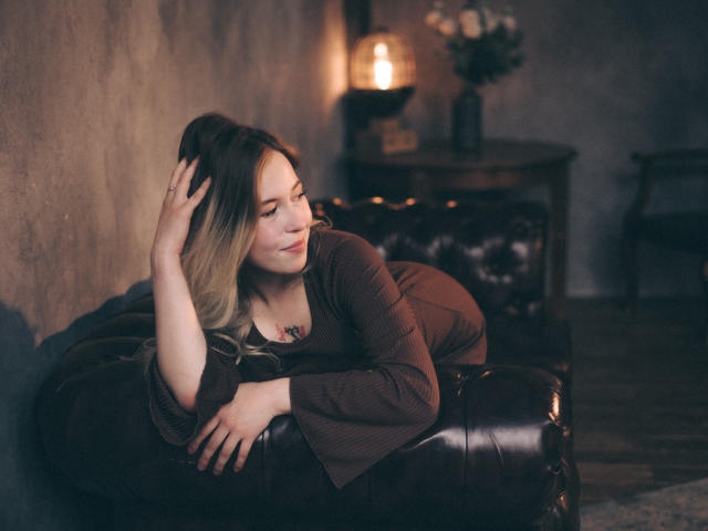 LanaRoselle met blond haar, een bruine jurk en een tatoeage, liggend op een donkerbruine leren bank, hand in haar haar, naar de zijkant kijkend, zacht licht, donkere kamer, ontspannen, sexy, kalm..